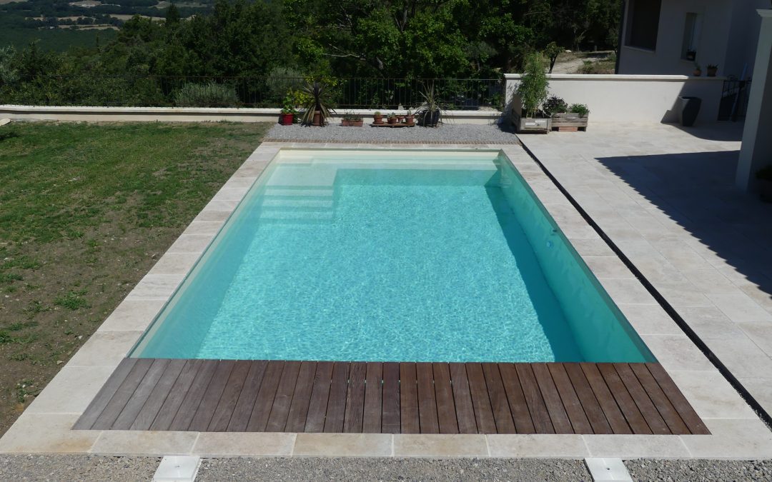 Piscine coque beige avec plage en pierre et en bois, sur un terrain en hauteur face aux collines