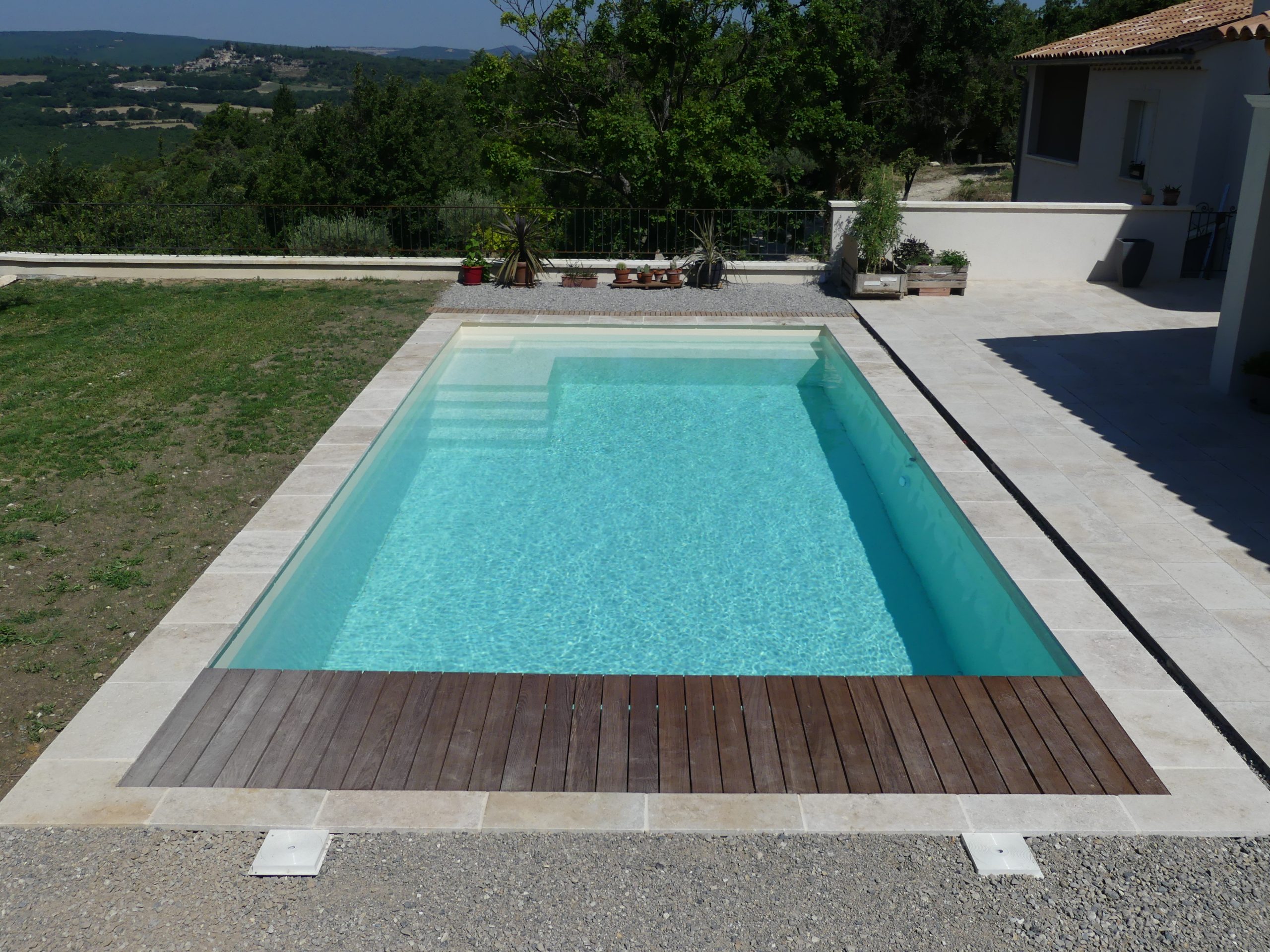 Piscine coque beige avec plage en pierre et en bois, sur un terrain en hauteur face aux collines